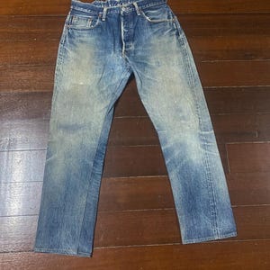 Op de afbeelding: Verbleekte blauwe spijkerbroek met een knoopsluiting en een klassiek vijf-zakken ontwerp. De jeans vertonen tekenen van slijtage en vervaging, met lichtere vlekken op de dijen en knieën. De jeans liggen plat op een houten oppervlak.