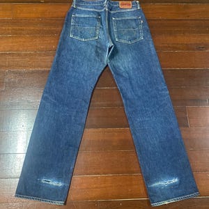 May include: Dark blue denim jeans with a classic five-pocket design. The jeans feature a faded wash and distressed details near the knees. A red leather patch with the brand name is visible on the back waistband.