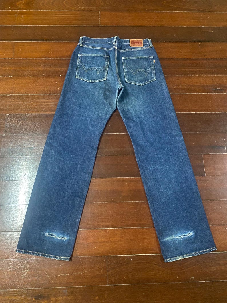 May include: Dark blue denim jeans with a classic five-pocket design. The jeans feature a faded wash and distressed details near the knees. A red leather patch with the brand name is visible on the back waistband.