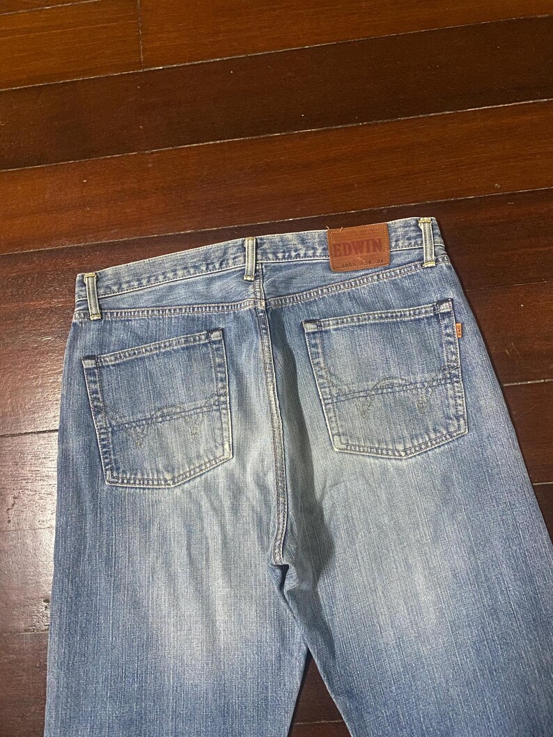 May include: Light blue denim jeans with a classic five-pocket design. The back of the jeans features two pockets and a brown leather patch with the brand name "EDWIN". The jeans are made of a durable denim fabric.