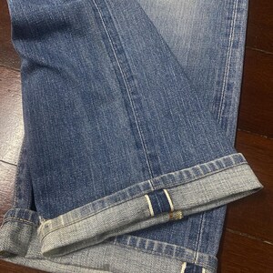 May include: Close-up of blue denim jeans with folded cuffs. The jeans have a classic five-pocket design and a faded wash. The cuffs are turned up, revealing the selvedge edge with a white and red stripe.