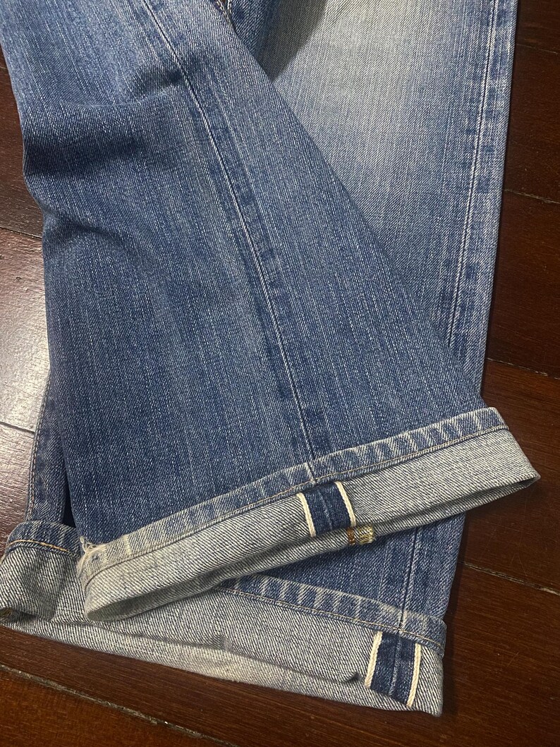 May include: Close-up of blue denim jeans with folded cuffs. The jeans have a classic five-pocket design and a faded wash. The cuffs are turned up, revealing the selvedge edge with a white and red stripe.