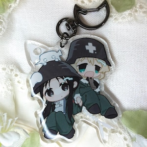 Könnte beinhalten: Ein klarer Acryl-Schlüsselanhänger mit zwei Anime-Figuren in grünen Uniformen und schwarzen Hüten. Eine Figur hat lange schwarze Haare, die andere blonde Haare. Der Schlüsselanhänger hat einen schwarzen katzenförmigen Verschluss und einen silbernen Ring.