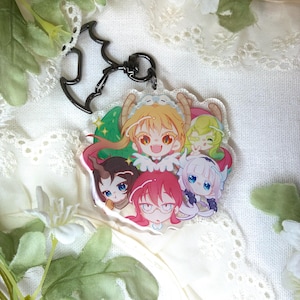Puede incluir: Un llavero acrílico transparente con un grupo de personajes de estilo anime con cabello y expresiones coloridas. El llavero tiene un cierre de metal negro y un accesorio en forma de murciélago. El fondo es una tela blanca suave con detalles florales.