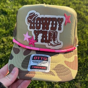 Op de afbeelding: Een beige trucker-pet met een camouflagepatroon aan de voorkant. De pet heeft een roze kralenrand en een bruin patch met de tekst "HOWDY Y'ALL" met een cowboyhoed- en laarsendesign. De pet heeft ook twee roze sterren aan de zijkant.