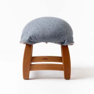 May include: A small, round stool with a light blue fabric seat and wooden legs. The seat is covered in a textured, woven fabric. The wooden legs are a light brown color and have a curved design. The word "TUFFET" is printed on the front of the stool.