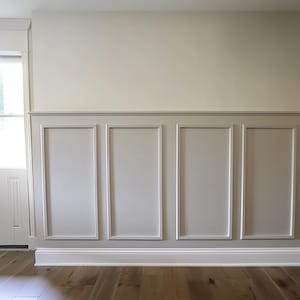 May include: A white wall with wainscoting, featuring four rectangular panels with raised edges. The wall is painted a light beige color and the panels are a slightly darker shade of beige. The wainscoting is finished with a white baseboard.