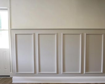May include: A white wall with wainscoting, featuring four rectangular panels with raised edges. The wall is painted a light beige color and the panels are a slightly darker shade of beige. The wainscoting is finished with a white baseboard.