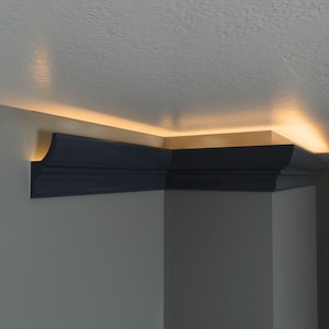Könnte beinhalten: Eine dunkel anthrazitfarbene Deckenleiste, die entlang der Oberkante einer Wand am Übergang zur Decke angebracht ist. Die Leiste hat ein geschwungenes Profil und wird von hinten durch ein warmes Licht beleuchtet, das auf die Decke strahlt.