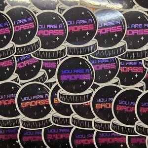 May include: A pile of black and white stickers with a glitter finish. Each sticker features a crystal ball with the text "You are a badass" in pink, purple, or blue.