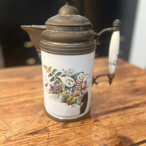 May include: A vintage brass and porcelain pitcher with a floral design. The pitcher has a white porcelain body with a floral pattern in shades of pink, purple, green, and yellow. The top and handle are made of brass.