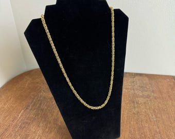 May include: A gold-colored chain necklace displayed on a black velvet jewelry stand. The necklace has a woven rope design. The stand is angled, and the background is a neutral color.