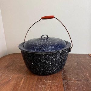 May include: A speckled black and blue enamelware pot with a lid and a wire handle with a red wooden grip. The pot sits on a wooden surface. The pot is a vintage kitchenware item.