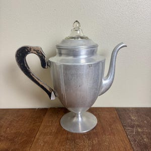 May include: Vintage aluminum teapot with a clear glass lid and a black and brown handle. The teapot has a curved spout and sits on a pedestal base. The body of the teapot has vertical lines.