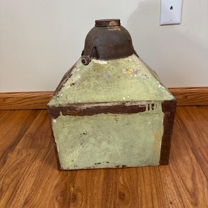 May include: An antique metal container with a light green, weathered exterior and a dark, rusty brown trim. The top is a rounded, darker metal with a small, circular lid. The container has a unique, geometric shape and shows signs of age.