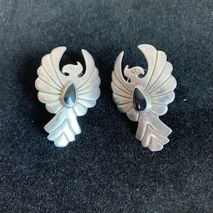 May include: A pair of silver-toned earrings, each shaped like a stylized bird with outstretched wings. The earrings feature a black, teardrop-shaped stone at the center of the bird's body. The wings have a detailed, fanned design.