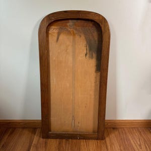 May include: A vintage wooden framed piece with a light-colored wooden interior. The frame is a dark brown color with a rounded top. The interior has a vertical seam down the middle and some dark staining on the right side.