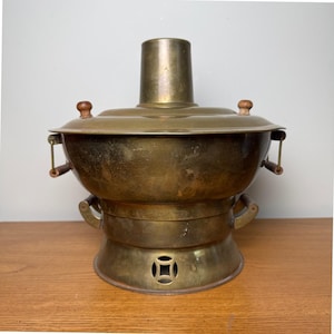 May include: A vintage brass hot pot with a central chimney and wooden handles. The pot has a round base with a decorative square cut-out and a lid with wooden knobs. The pot is sitting on a wooden surface.
