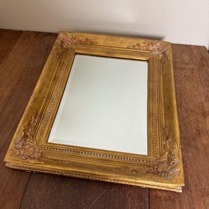 May include: A rectangular mirror with a gold-colored ornate frame. The frame features detailed carvings at the corners and along the edges. The mirror is set on a wooden surface.
