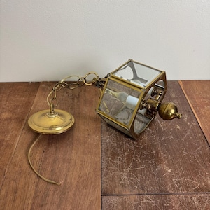 May include: Vintage brass pendant light fixture with a geometric glass enclosure. The fixture has a chain and a round ceiling plate. The light bulb is visible inside the glass enclosure. The fixture is resting on a wooden surface.