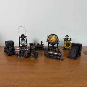 May include: A collection of miniature metal pencil sharpeners featuring a vintage car, a lantern, a motorcycle, two steam locomotives, a globe on a stand, a clock with a yellow face, and a box camera, all finished in a dark bronze metallic tone.