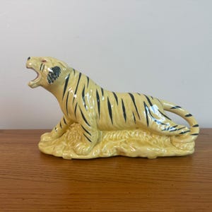 May include: A ceramic tiger figurine in a crouching pose, with an open mouth. The tiger has a cream-colored body with black stripes and red eyes. The figurine is resting on a textured, cream-colored base.