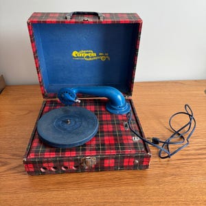 May include: Vintage portable record player with a red and black plaid exterior. The interior is blue, and it has a blue telephone-style arm and a black power cord. The brand name "Carron" is visible inside the lid.
