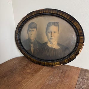 May include: Oval antique portrait of two people in sepia tones, framed in a dark wood frame with gold accents. The frame shows signs of age with some chipped areas. The subjects are dressed in formal attire.