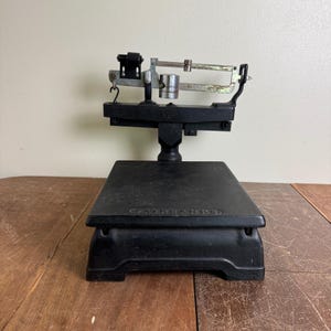 May include: An antique black Fairbanks scale with a rectangular base and a balance beam. The scale has a platform for weighing items and a beam with measurement markers. The scale is made of metal and sits on a wooden surface.