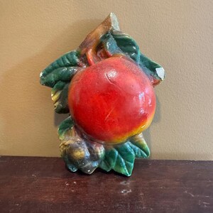 May include: A ceramic wall hanging featuring a red apple with green leaves and a brown stem. The apple has yellow and orange highlights. The leaves are a dark green color. The piece is likely vintage.