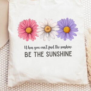 Könnte beinhalten: Eine weiße Stofftasche aus Segeltuch mit einer Grafik von drei Gänseblümchen in Rosa, Weiß und Violett. Unter den Blumen steht der Text: When you can't find the sunshine BE THE SUNSHINE. Die Tasche verfügt über zwei Tragegriffe.