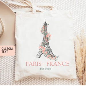 Könnte beinhalten: Eine weiße Canvas-Tragetasche mit einer Schwarzweiß-Illustration des Eiffelturms, der mit rosa Blumen verziert ist. Der Text "PARIS - FRANCE EST. 2025" ist unterhalb der Illustration aufgedruckt.