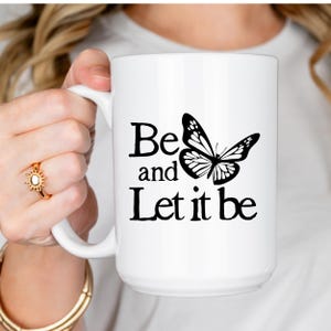 Könnte beinhalten: Ein weißer Keramikbecher mit einer schwarzen Grafik eines Schmetterlings und dem Text "Be and Let it be" auf der Vorderseite. Der Becher hat eine zylindrische Standardform mit einem geschwungenen Henkel an der Seite für eine klare Sicht.
