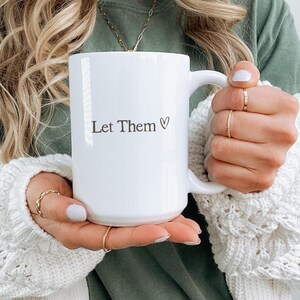 K&ouml;nnte beinhalten: Wei&szlig;er Keramikbecher mit dem Text "Let Them" und einem Herzsymbol. Der Becher wird von einer Person gehalten, die einen wei&szlig;en Pullover tr&auml;gt. Der Becher hat einen gro&szlig;en Henkel.