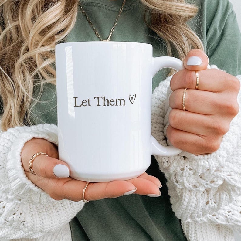 K&ouml;nnte beinhalten: Wei&szlig;er Keramikbecher mit dem Text "Let Them" und einem Herzsymbol. Der Becher wird von einer Person gehalten, die einen wei&szlig;en Pullover tr&auml;gt. Der Becher hat einen gro&szlig;en Henkel.