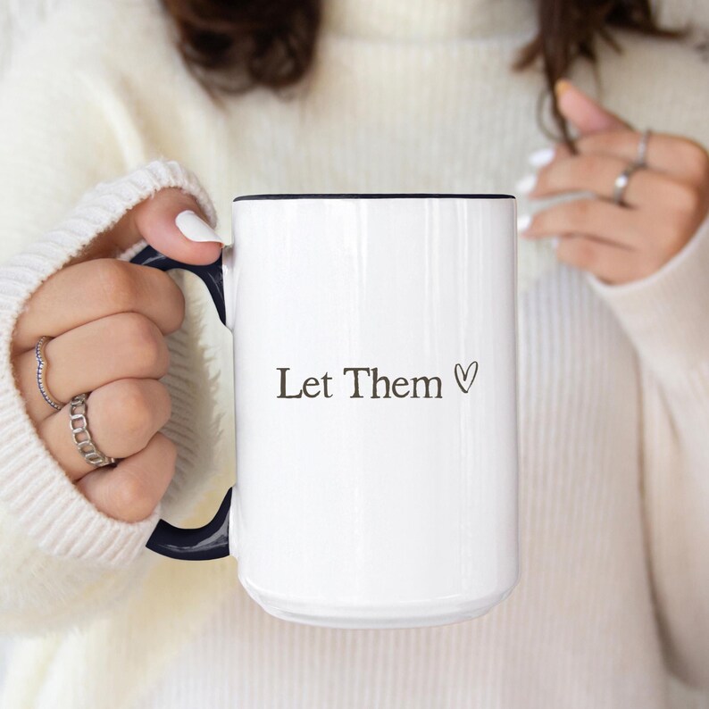 K&ouml;nnte beinhalten: Wei&szlig;er Keramikbecher mit schwarzem Rand und Henkel. Der Becher tr&auml;gt den Text "Let Them" mit einem Herzsymbol. Der Becher wird von einer Person gehalten, die Ringe und einen wei&szlig;en Pullover tr&auml;gt.