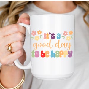 Könnte beinhalten: Ein weißer Keramikbecher mit dem bunten Schriftzug It's a good day to be happy in einer Retro-Schriftart. Das Design enthält kleine pastellfarbene Blumen in Rosa, Gelb und Orange, die den Text umgeben. Der Becher hat eine glatte Oberfläche.