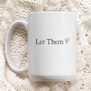K&ouml;nnte beinhalten: Eine wei&szlig;e Keramiktasse mit gebogenem Henkel. Auf der Tasse steht der Schriftzug "Let Them" in dunkelgrauer Schrift, gefolgt von einem kleinen Herzsymbol. Die Tasse steht vor einem strukturierten, cremefarbenen Hintergrund.
