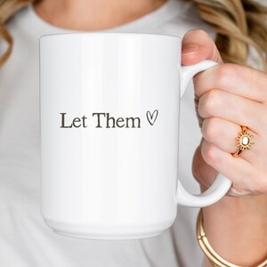 K&ouml;nnte beinhalten: Eine wei&szlig;e Keramik-Tasse mit dem Text "Let Them" und einem schwarzen Herzsymbol. Die Tasse hat einen gro&szlig;en Henkel und eine glatte, gl&auml;nzende Oberfl&auml;che, ideal f&uuml;r Hei&szlig;getr&auml;nke.