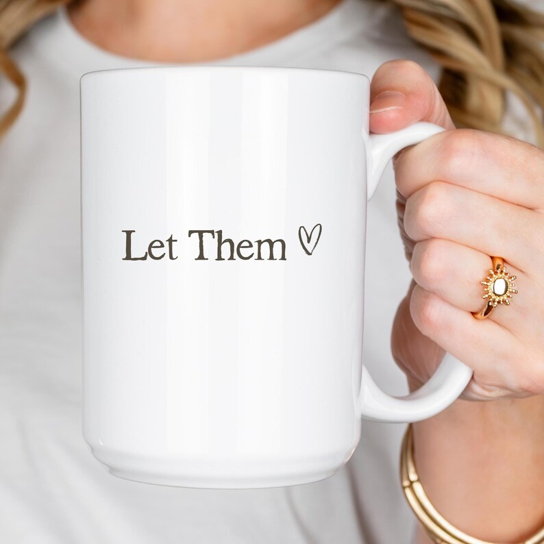 K&ouml;nnte beinhalten: Eine wei&szlig;e Keramik-Tasse mit dem Text "Let Them" und einem schwarzen Herzsymbol. Die Tasse hat einen gro&szlig;en Henkel und eine glatte, gl&auml;nzende Oberfl&auml;che, ideal f&uuml;r Hei&szlig;getr&auml;nke.