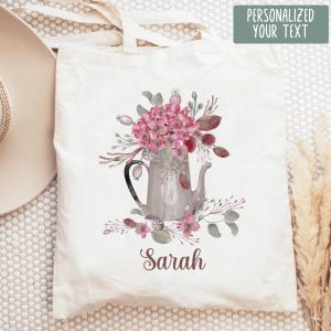 Könnte beinhalten: Eine cremefarbene Stofftasche aus Segeltuch mit einer Aquarellillustration einer grauen Teekanne, die mit rosa Hortensienblüten und grünem Blattwerk gefüllt ist. Der Name Sarah ist in einer dunkelbraunen Schreibschrift unter dem Motiv gedruckt.
