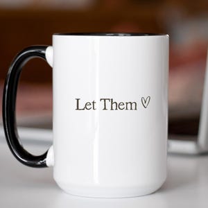 K&ouml;nnte beinhalten: Wei&szlig;er Keramikbecher mit schwarzem Henkel und Rand. Der Becher tr&auml;gt den Text "Let Them" mit einem Herzsymbol. Der Becher steht auf einer wei&szlig;en Oberfl&auml;che, mit verschwommenem Hintergrund.