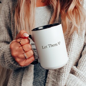 K&ouml;nnte beinhalten: Wei&szlig;er Keramikbecher mit schwarzem Innenraum und Henkel, mit dem Text "Let Them" mit einem Herzsymbol. Der Becher wird von einer Person gehalten, die einen grauen Pullover tr&auml;gt. Der Becher hat eine Standardgr&ouml;&szlig;e.