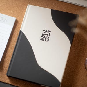 Könnte beinhalten: Ein schwarz-weißes Notizbuch mit den Zahlen "25 26" auf dem Einband, neben einem "To Do" Notizblock und einer Brille. Das Notizbuch hat einen weißen Einband mit einem schwarzen Wellenmuster. Der Notizblock ist weiß mit einer grauen Überschrift.