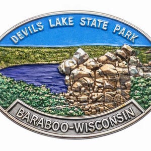 May include: An oval metal belt buckle featuring a raised relief landscape of Devils Lake State Park. The design includes a blue sky, a dark blue lake, and textured rock formations with green foliage. The text reads Devils Lake State Park and Baraboo-Wisconsin.