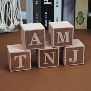 May include: Wooden alphabet blocks with light wood sides and dark wood faces. The blocks spell out the word "ANTJAM". The letters are in a simple, sans-serif font. The blocks are arranged on a brown surface.