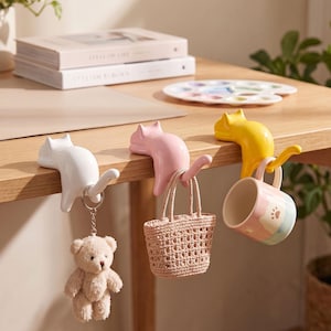 Peut inclure: Trois crochets en forme de chat, de couleurs blanche, rose et jaune, sont fixés au bord d'une table en bois. Le crochet blanc porte un porte-clés ours en peluche, le rose soutient un sac à main tressé et le jaune retient une petite tasse.