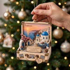 May include: A suitcase-shaped Christmas ornament featuring a 3D scene of Santorini with blue-domed buildings, a sunset sea view, and a plaque reading OLIVIA & HENRY SANTORINI HONEYMOON 2026. The exterior has travel stickers and a heart-shaped tag.