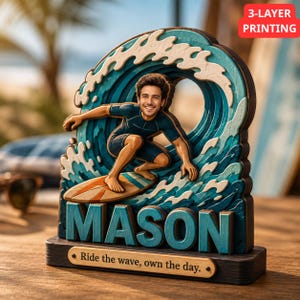 Puede incluir: Un recorte de madera de tres capas que muestra a un surfista sobre una ola con el nombre MASON en letras grandes de color azul verdoso. Una pequeña placa en la base dice Ride the wave, own the day. El diseño usa tonos de azul y blanco.