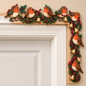 May include: A decorative door frame topper featuring seven brown and orange robins perched among green holly leaves, red berries, and gold bells. The ornament is arranged in an L-shape to fit the corner of a door frame, with red ribbons.
